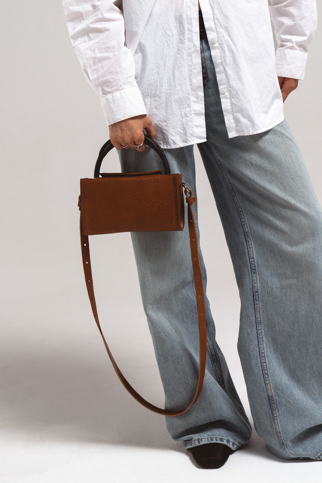 Brown leather AirPods Max crossbody case with long adjustable strap, handheld by model in white shirt and jeans