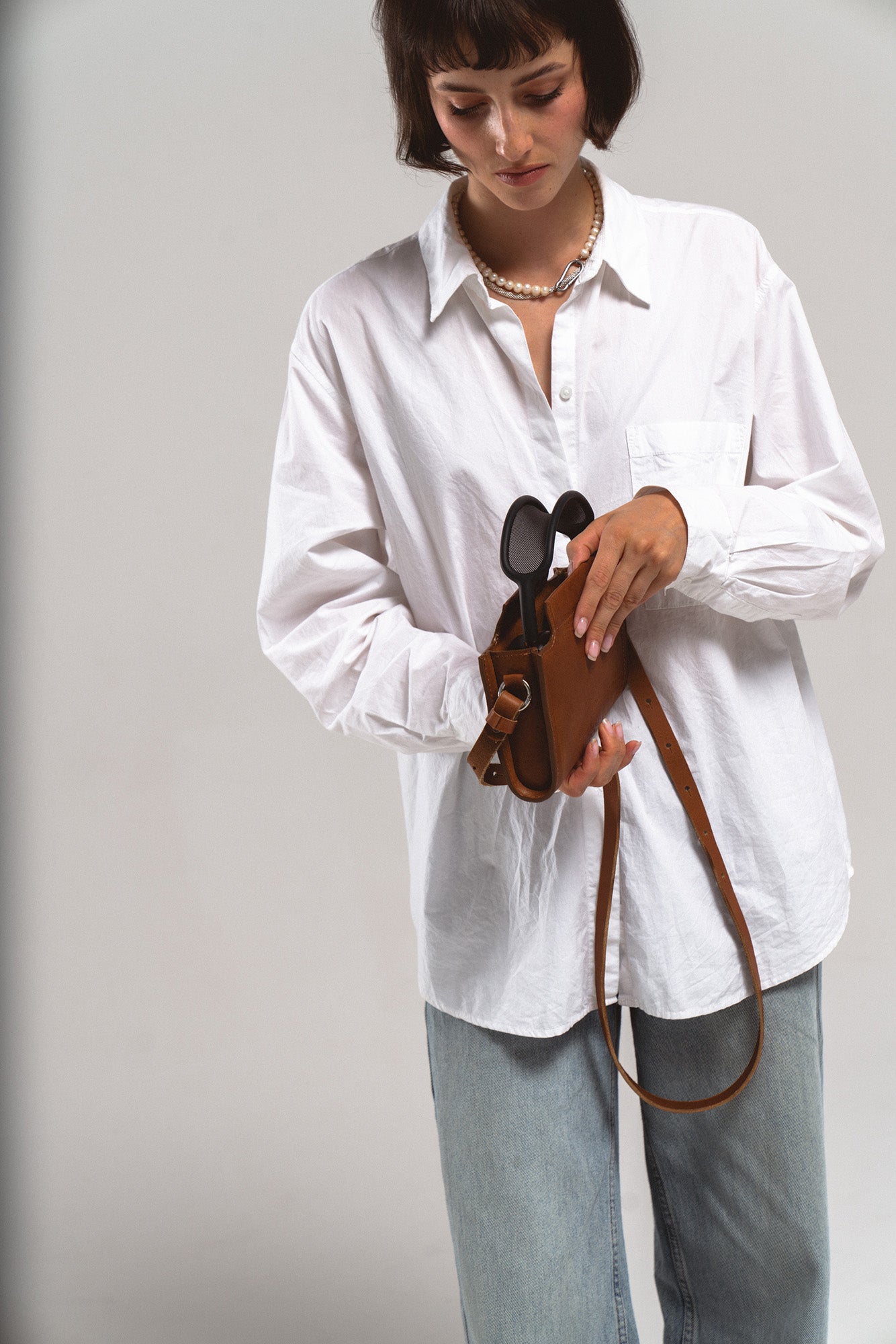 Model holding brown leather crossbody carrying case for Apple AirPods Max, placing headphones inside; adjustable strap and handle visible