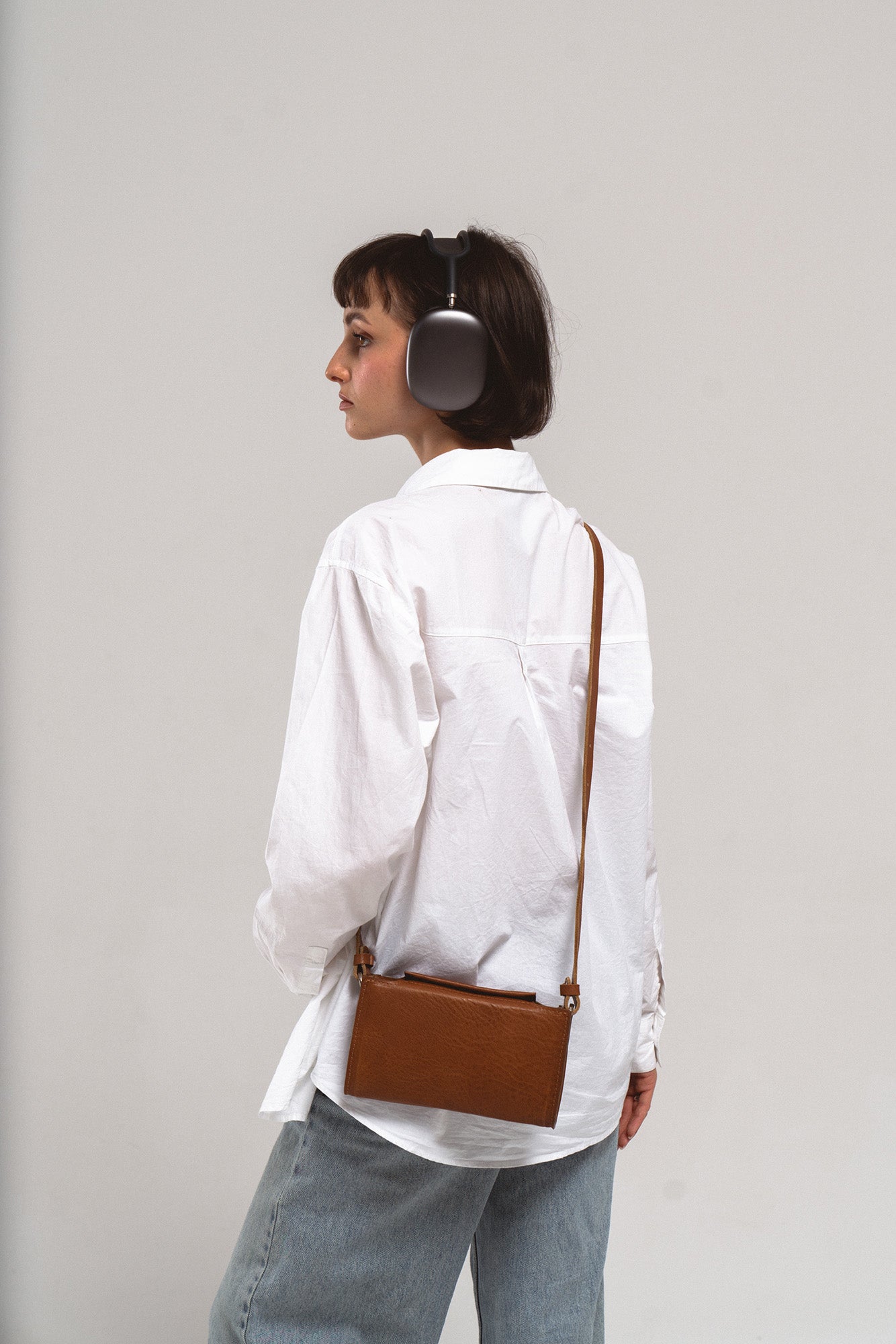Model wearing Apple AirPods Max with brown leather crossbody carrying case on long strap, back view in white shirt and jeans