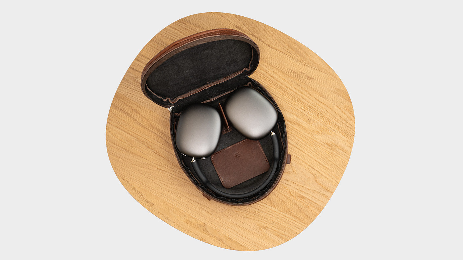 Top-down flat lay: AirPods Max inside open dark brown leather zip case with suede lining, on a round wooden tray.