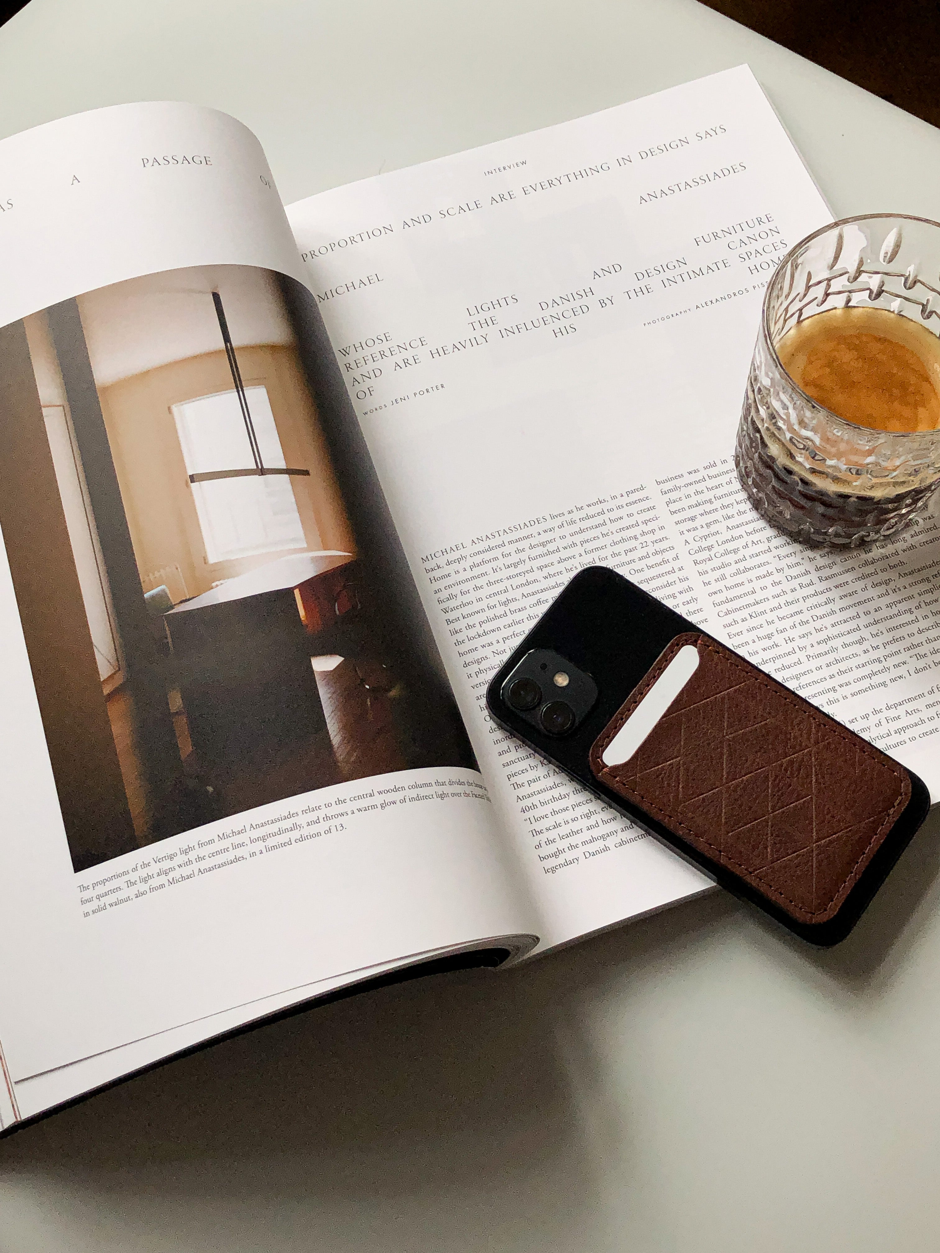 Brown leather card wallet attached to a smartphone resting on an open magazine next to a glass of coffee