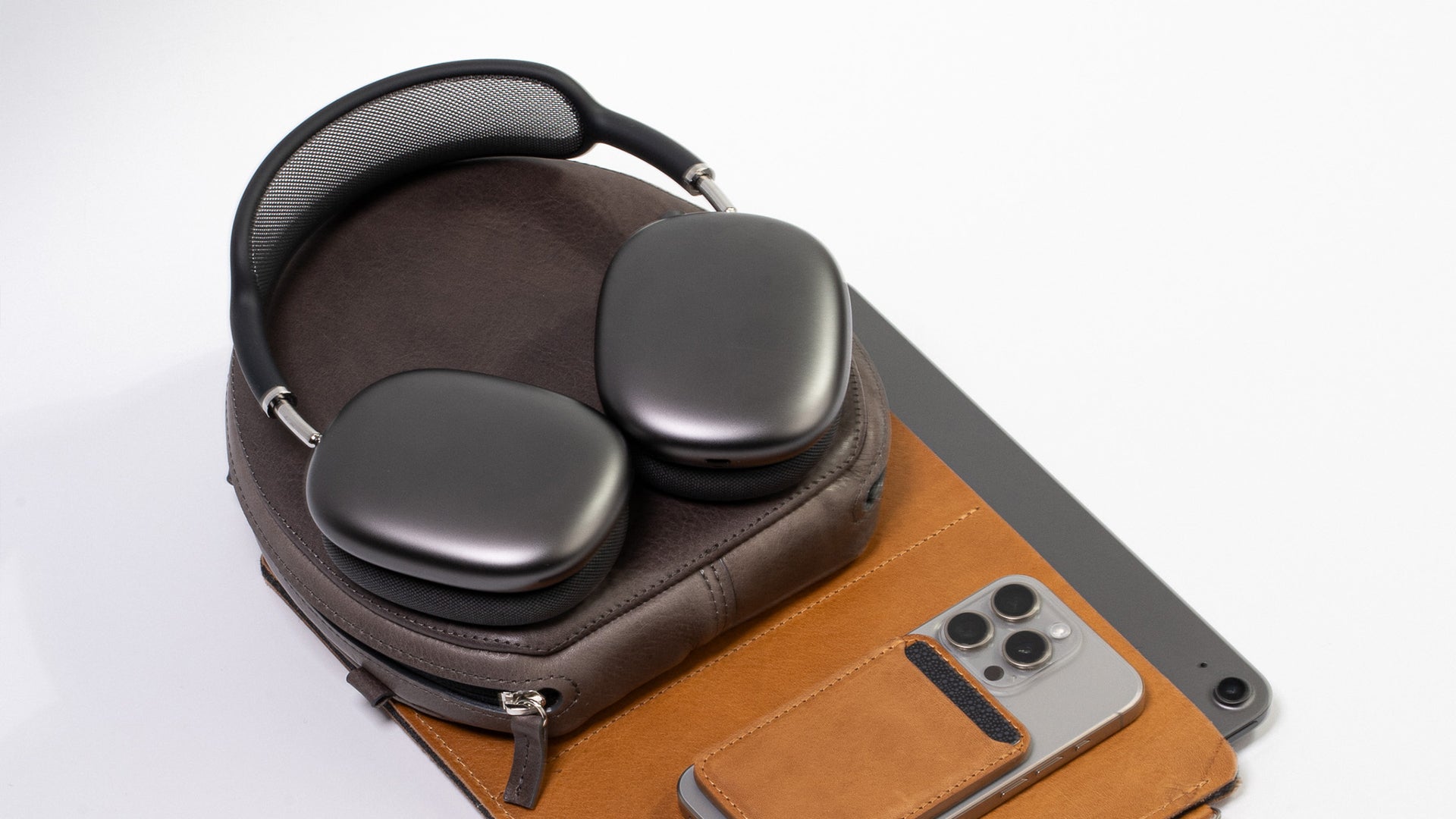 AirPods Max (space gray) resting on a closed dark-brown leather zip case, on a tan leather desk mat next to an iPhone Pro with triple-camera bump and a gray iPad (white background).