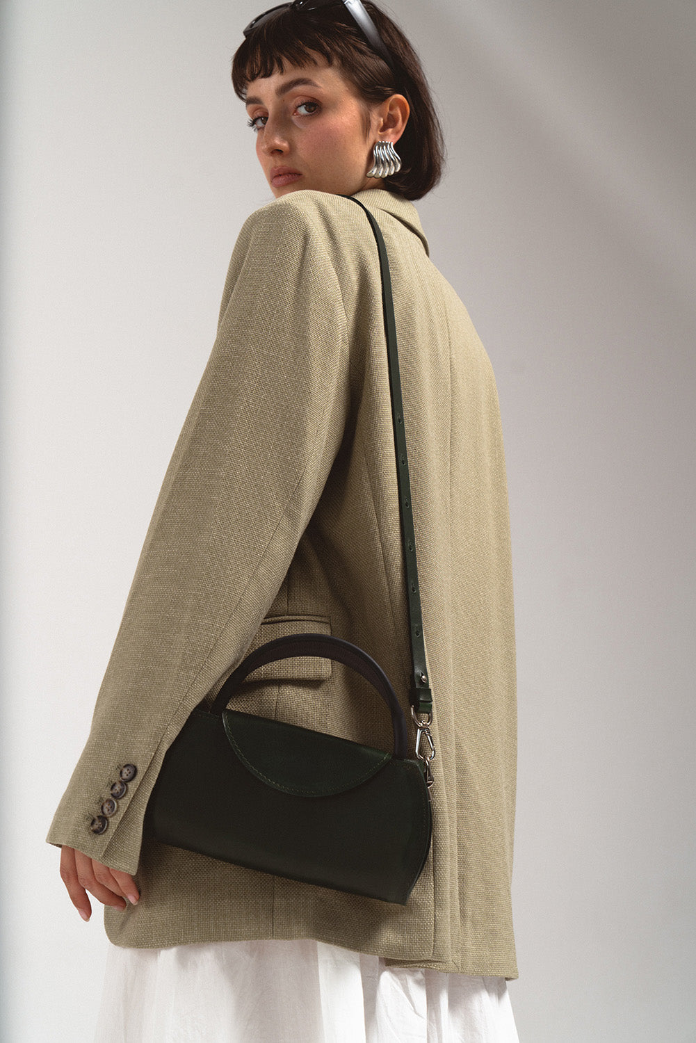 Dark green leather AirPods Max crossbody case worn on shoulder with top handle and long strap, studio product lifestyle