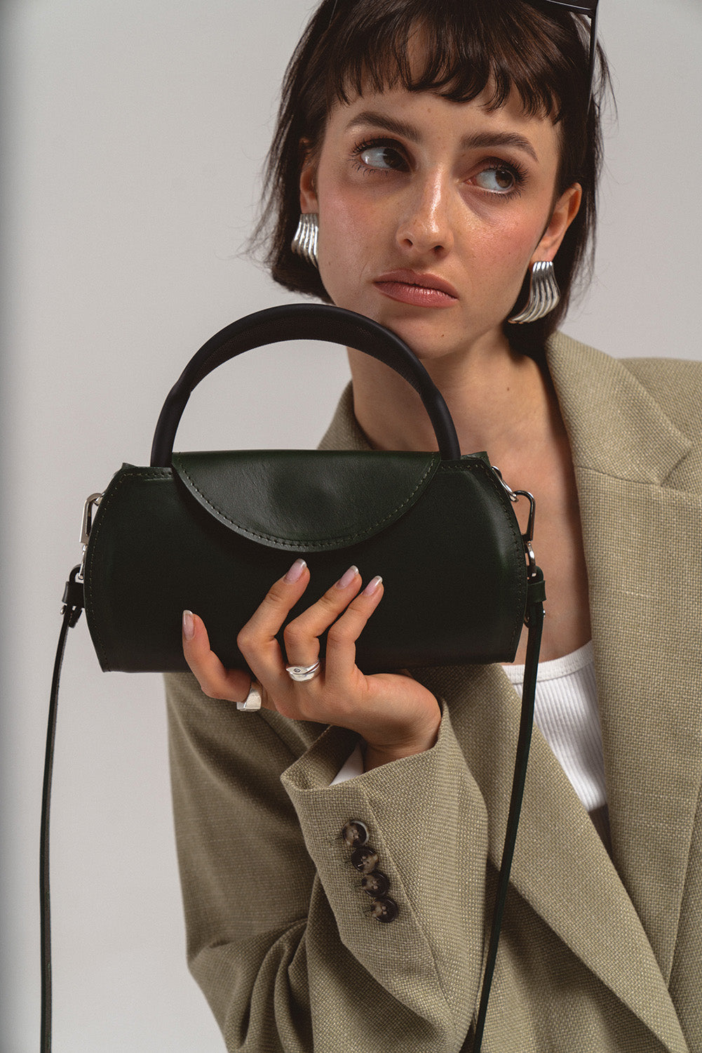 Woman holding a dark green leather AirPods Max case with top handle and shoulder strap, studio lifestyle photo