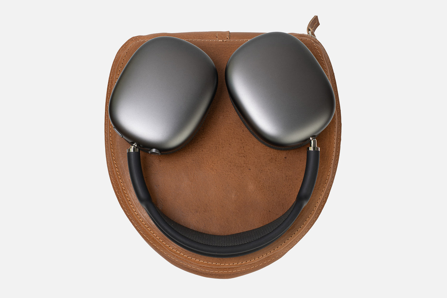 Top-down view of Apple AirPods Max resting on an open tan leather case insert (oval base) with stitched edge details, showing the headphones laid flat for storage/fit reference.