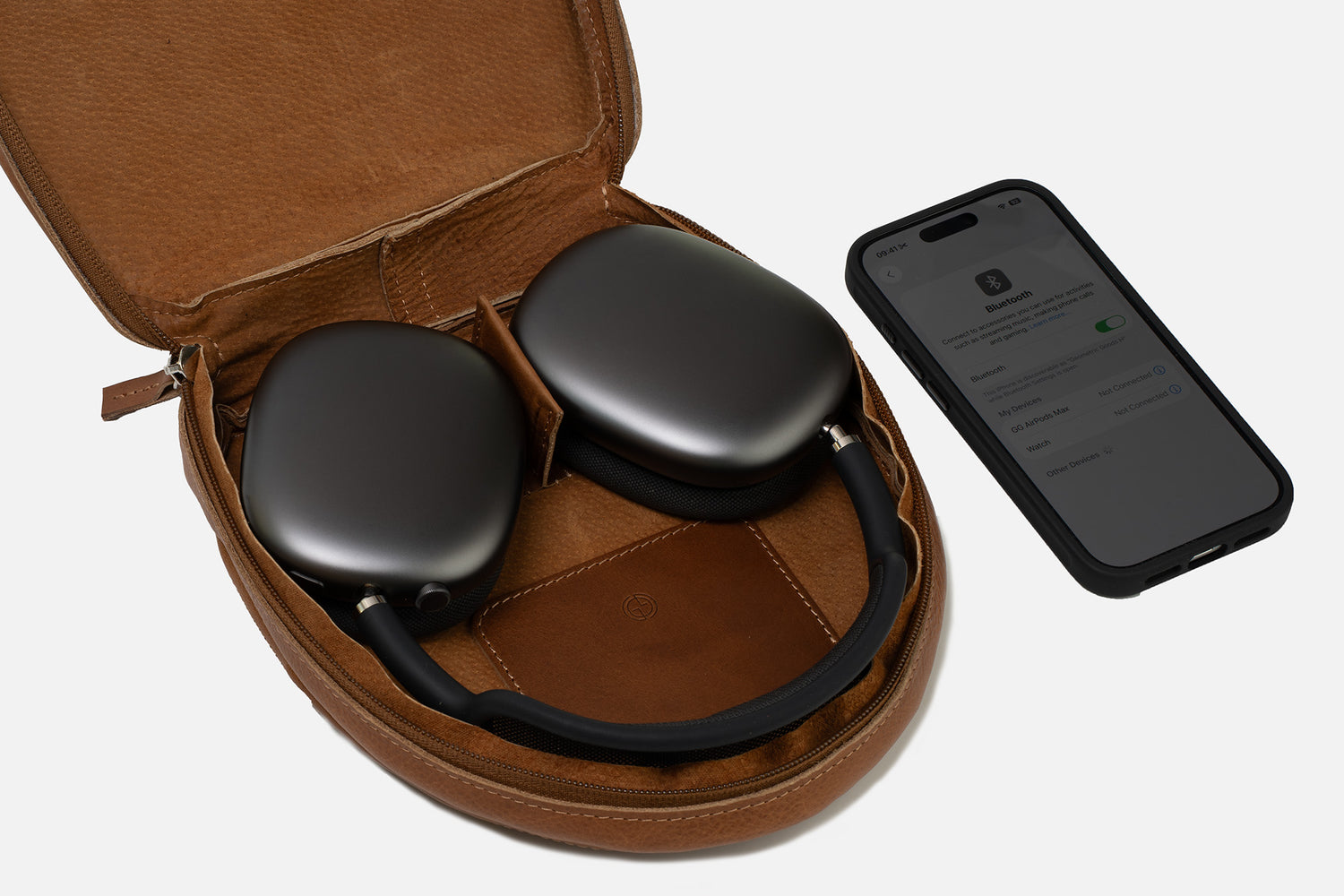 Open brown leather AirPods Max case showing the headphones stored flat inside with padded interior, central spacer panel, stitched pocket, and an iPhone Bluetooth screen beside it for scale.