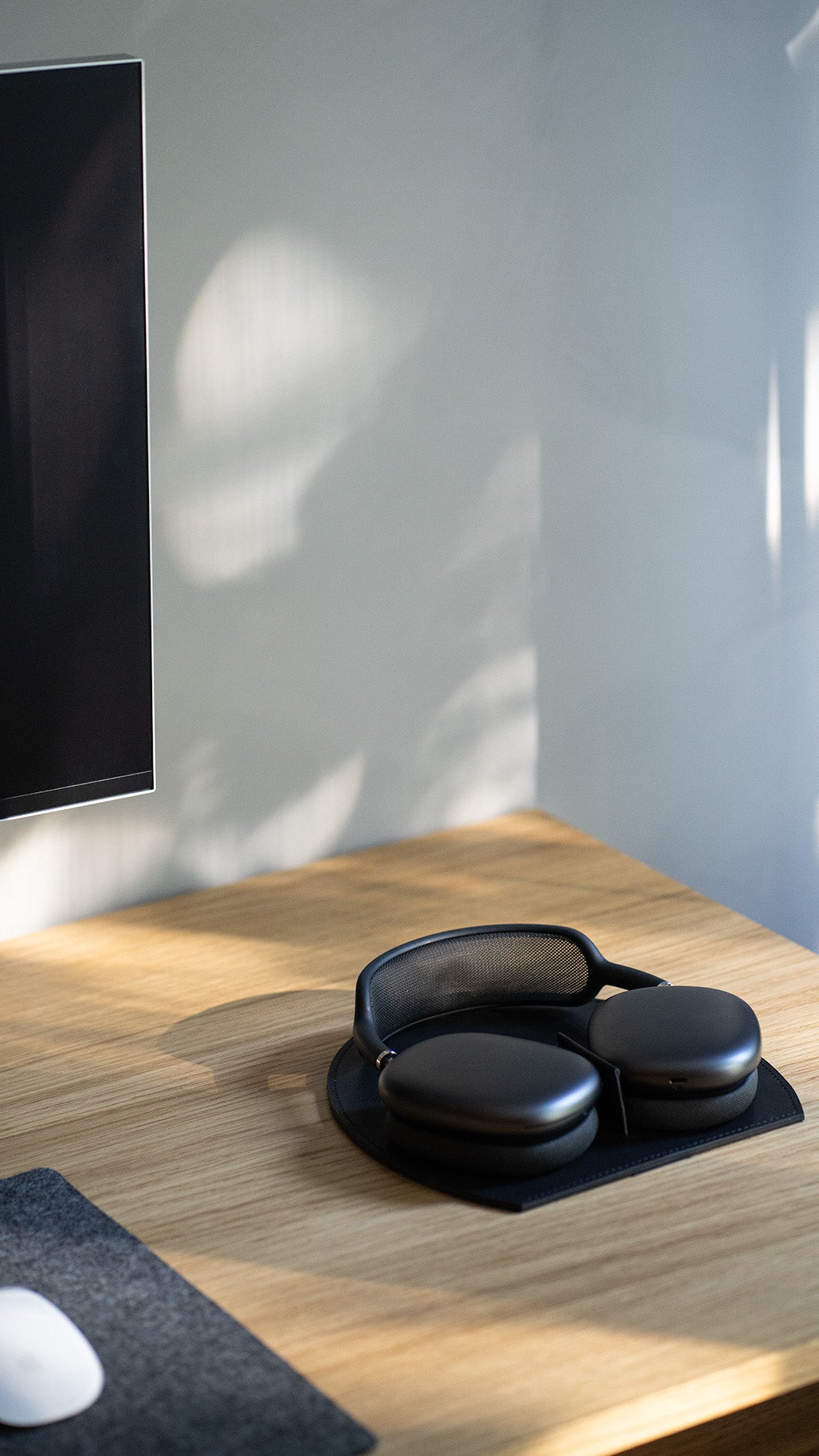 Black AirPods Max resting on a round black leather base with a center strap divider on a sunlit oak desk; soft window-shadow patterns on a light gray wall and a monitor edge visible at left.