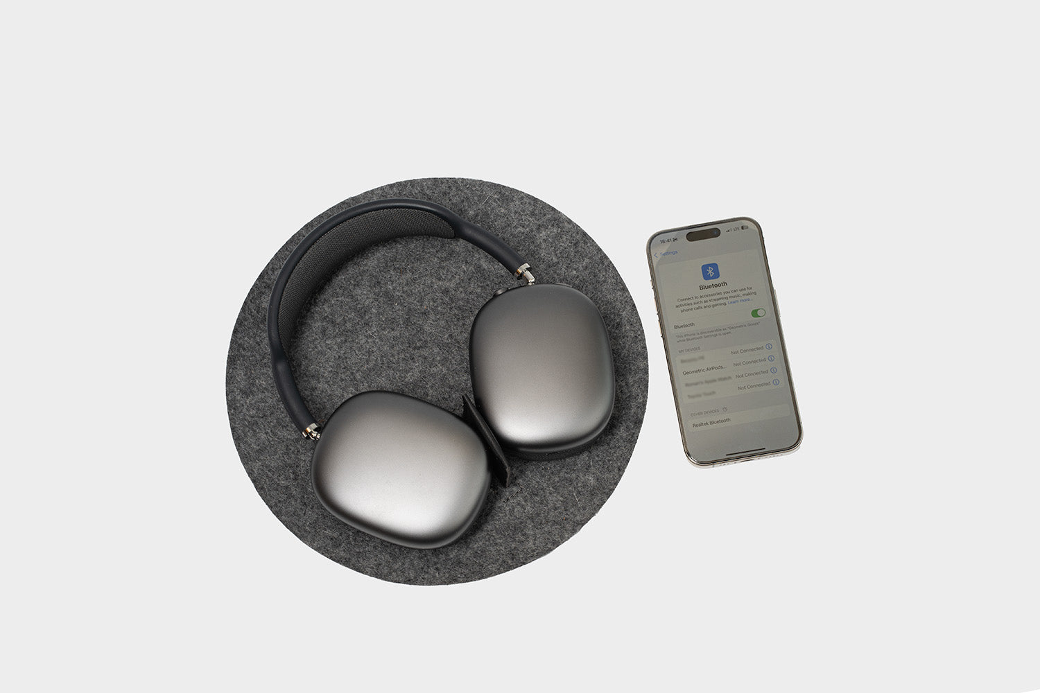 Top-down view of space-gray AirPods Max resting on a round dark gray wool-felt mat with a small black center divider, with a smartphone on the right showing the Bluetooth settings screen on a light gray background.