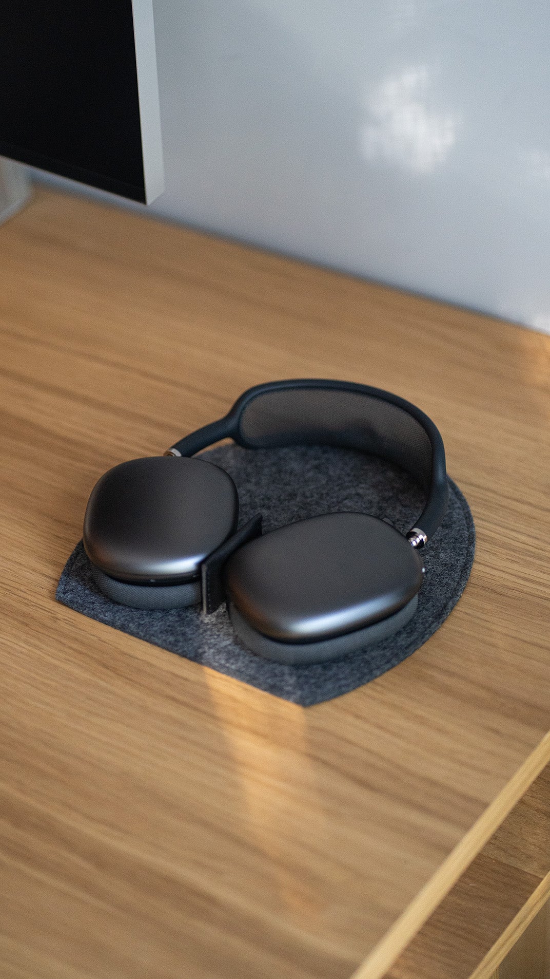 Space-gray AirPods Max lying flat on a dark gray wool-felt base with a black center divider; sunlit oak desk surface with a monitor corner at top left and a pale gray wall in the background.