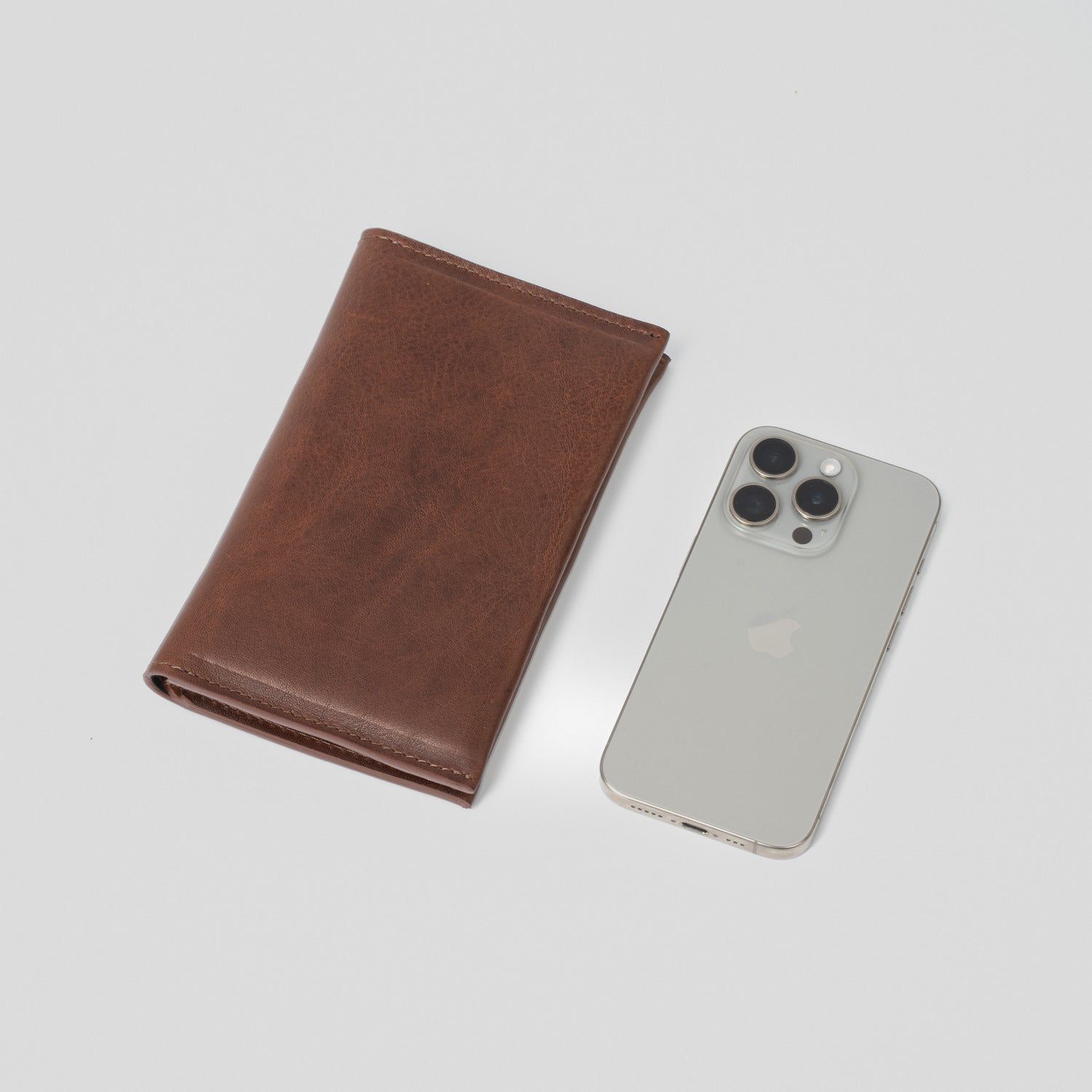 Dark brown leather passport travel wallet shown next to an iPhone for size comparison, photographed on a light background