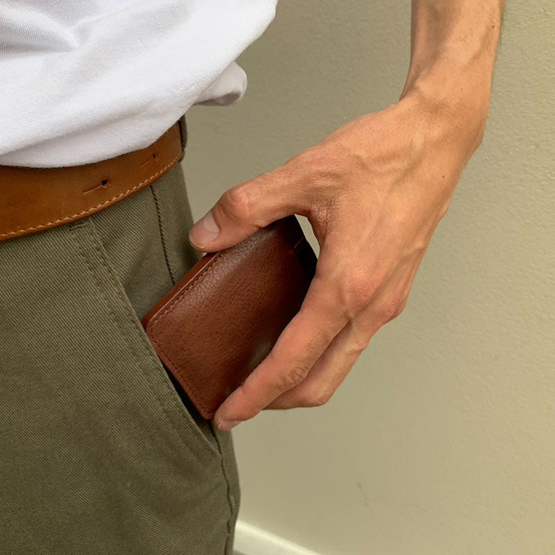 Hand sliding a slim brown leather wallet into a front pants pocket, showing compact everyday carry size