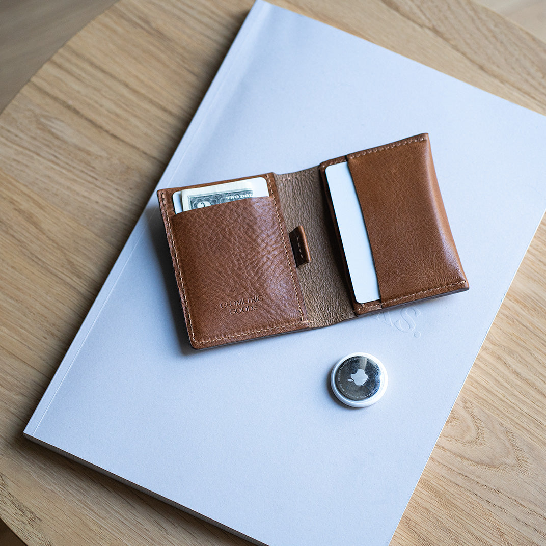 Open brown leather AirTag wallet with card slots and folded cash, placed on a book next to an Apple AirTag tracker on a wooden table