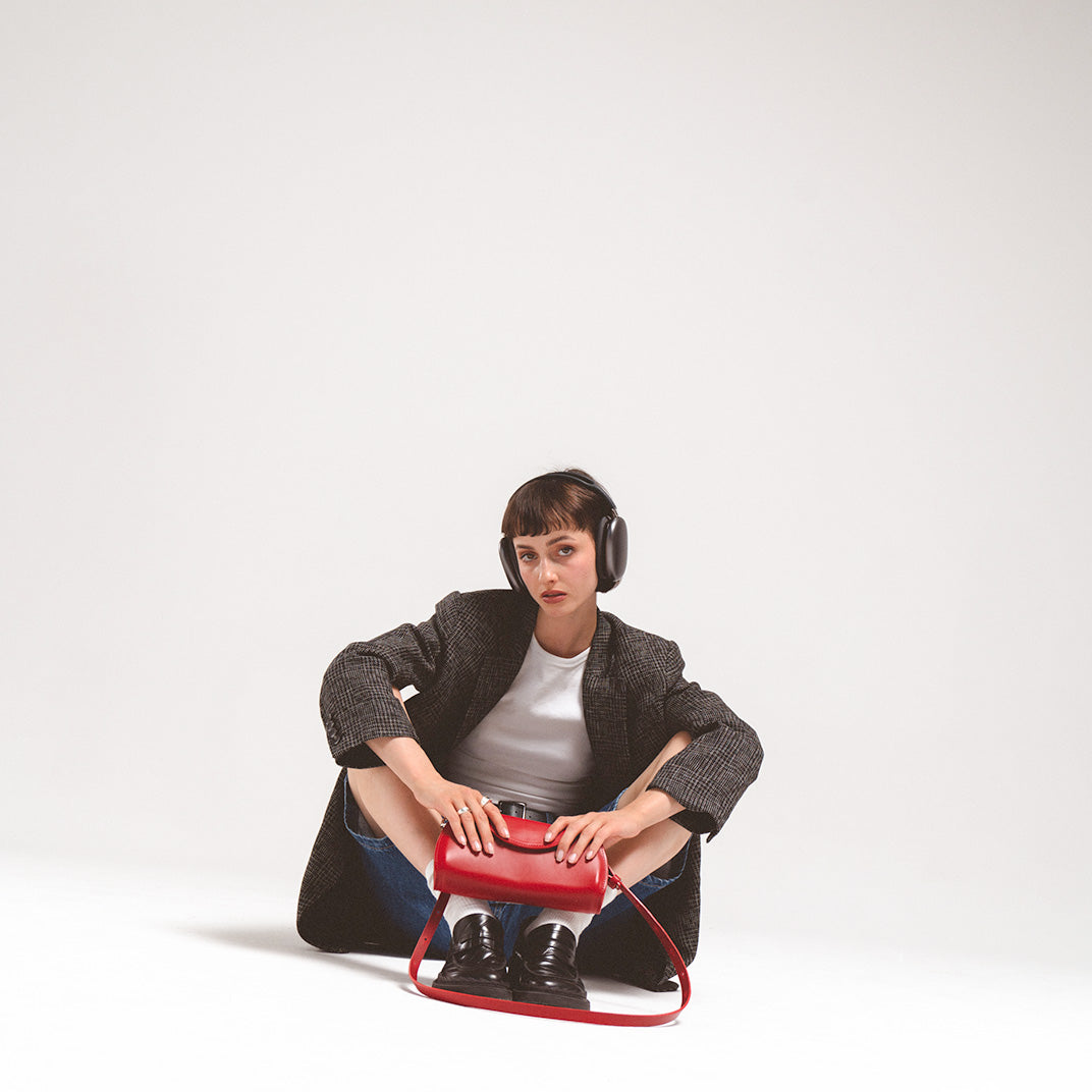 Leather case for AirPods Max in striking red, held by a seated young woman wearing headphones and a stylish blazer, showcasing a contemporary design