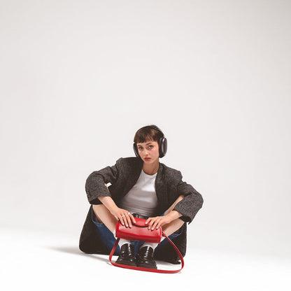 Leather case for AirPods Max in striking red, held by a seated young woman wearing headphones and a stylish blazer, showcasing a contemporary design