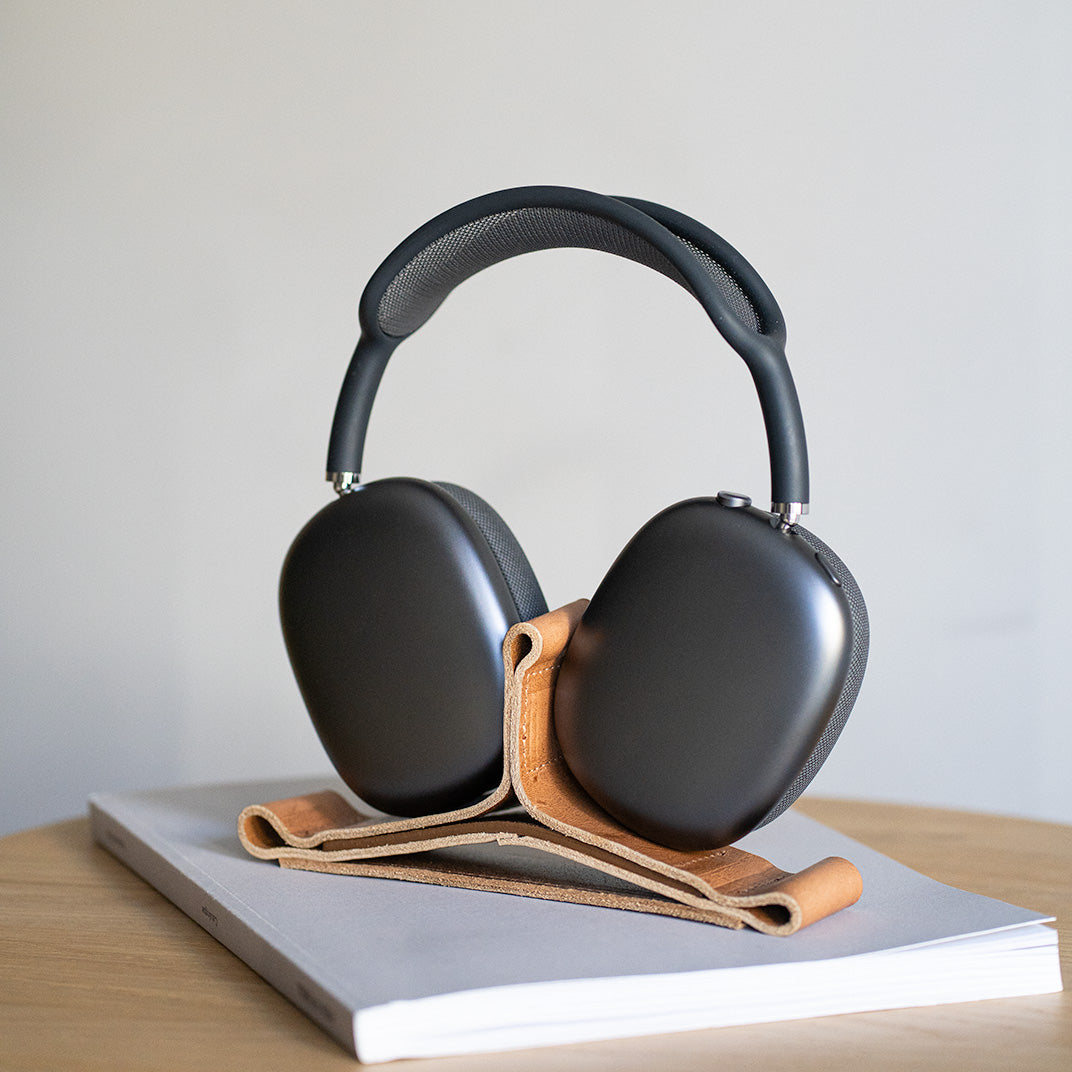Black AirPods Max headphones resting on a natural tan leather headphone stand on a book, on a wooden desk