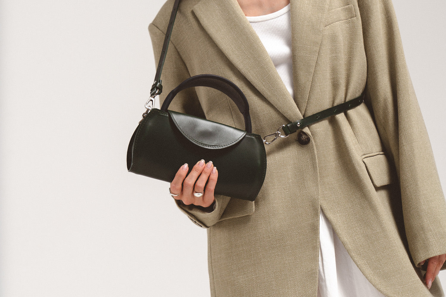 Model wearing a dark green leather AirPods Max case bag crossbody, with curved top handle, half-moon front flap, and silver clip hardware on the strap.