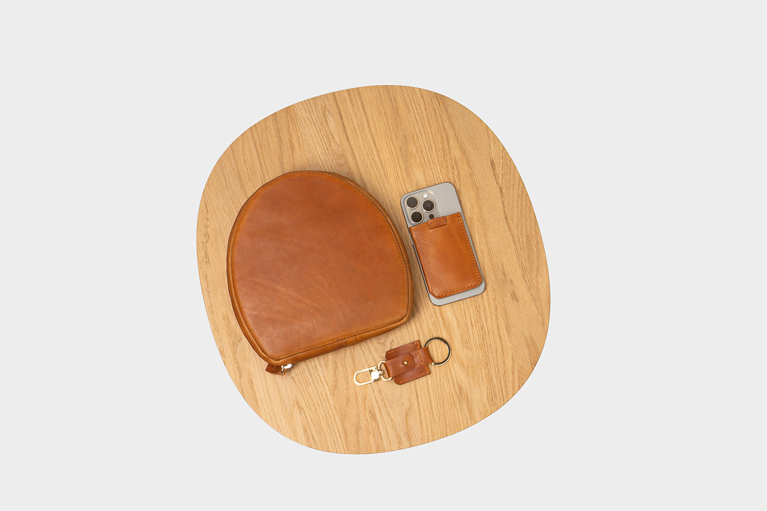 Flat lay on wooden tray: tan leather AirPods Max zip case with matching iPhone leather wallet sleeve and small leather key fob.