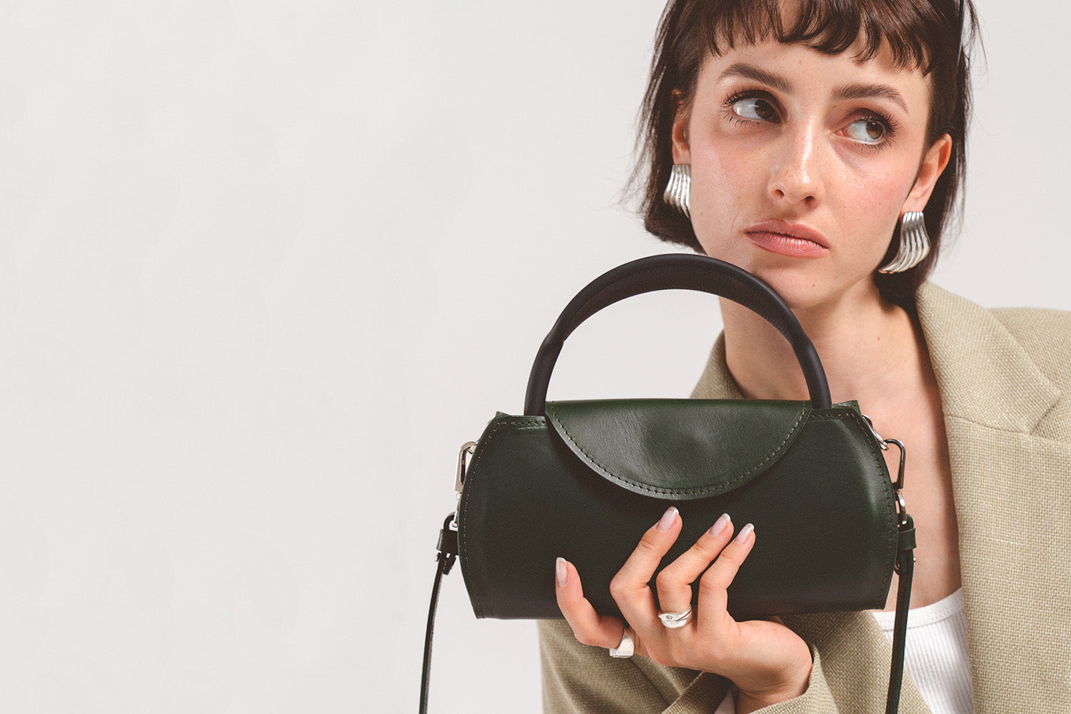 Model holding a dark green leather AirPods Max case bag with curved top handle and detachable shoulder strap, close-up studio portrait.