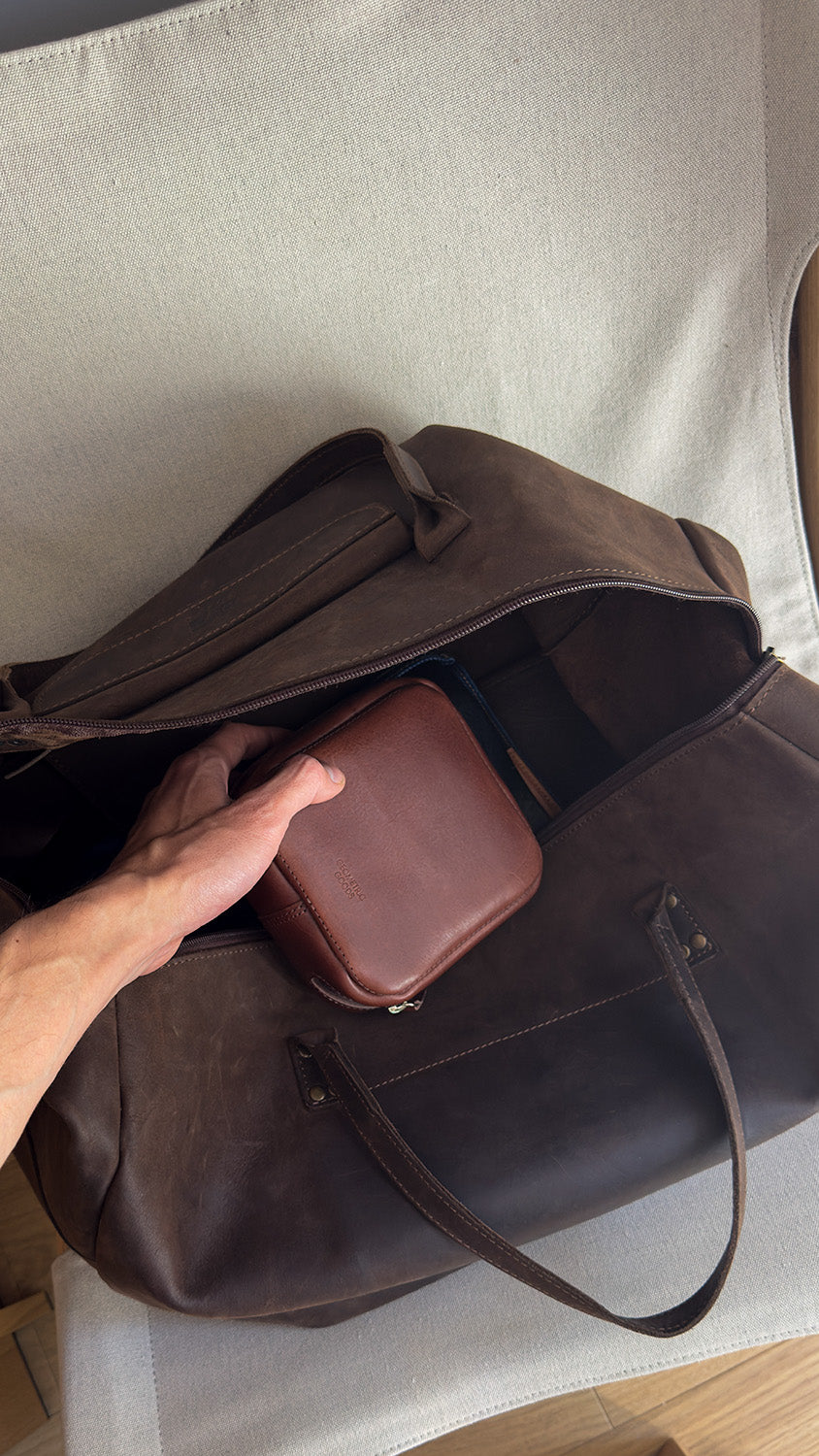 Hand placing a small brown leather case into an open leather duffle bag