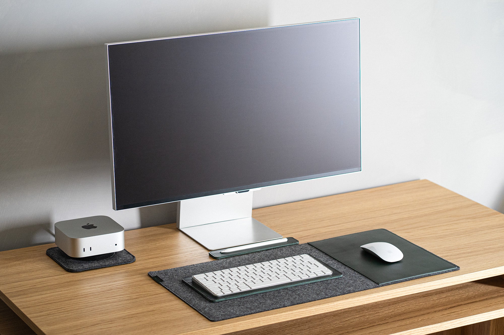 Minimal desk setup with Mac mini on a felt base, leather keyboard tray, and leather mouse pad on a wooden desk.