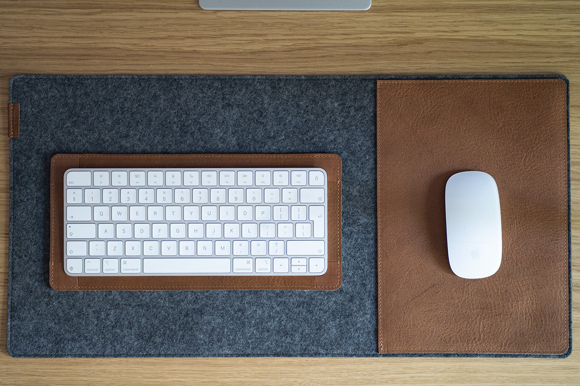Top-down view of a white wireless magic keyboard in a brown leather tray and a white mouse on a matching leather pad, set on a gray felt desk mat.
