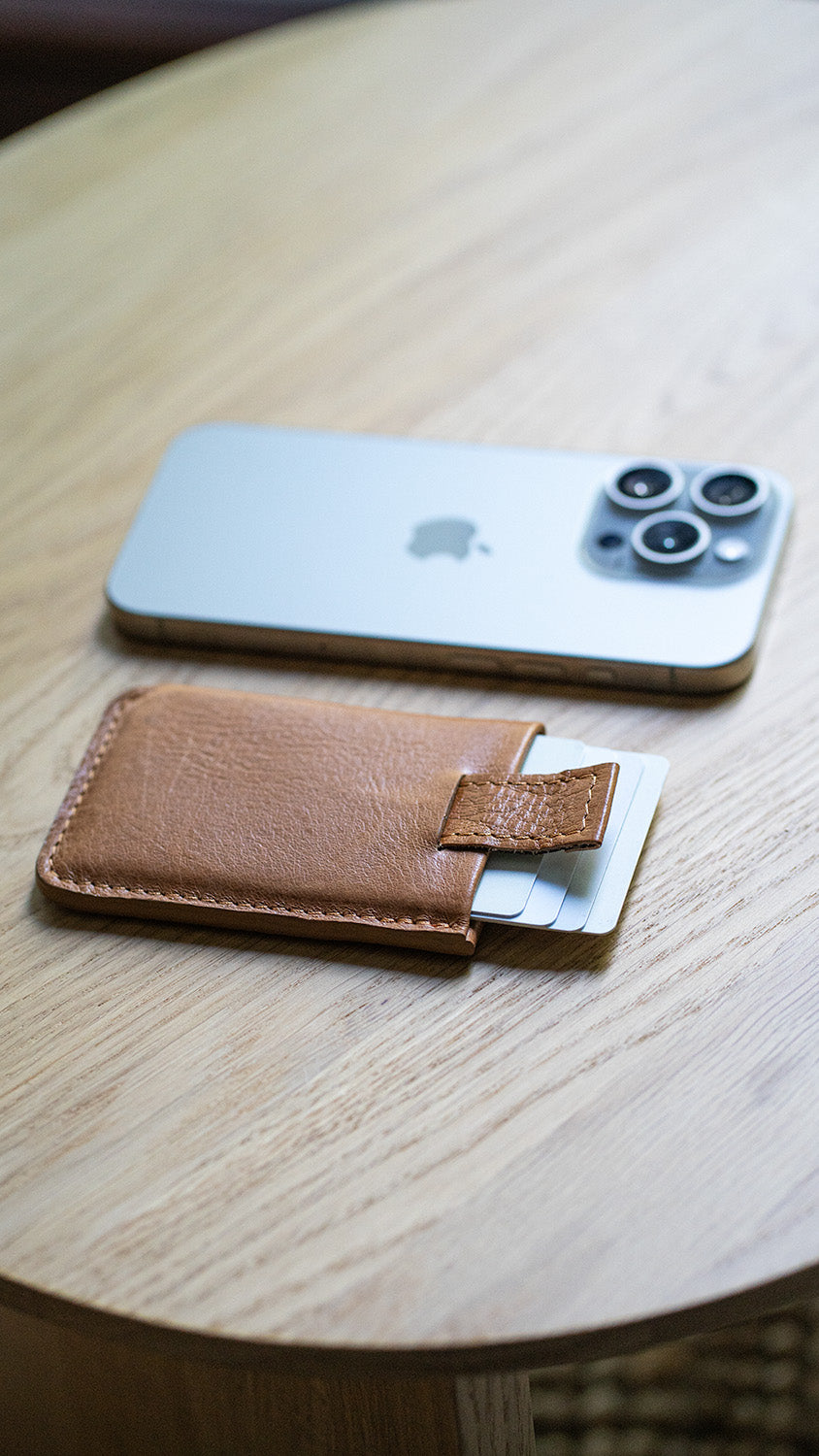 Brown leather card wallet with cards next to an iPhone on a wooden table.