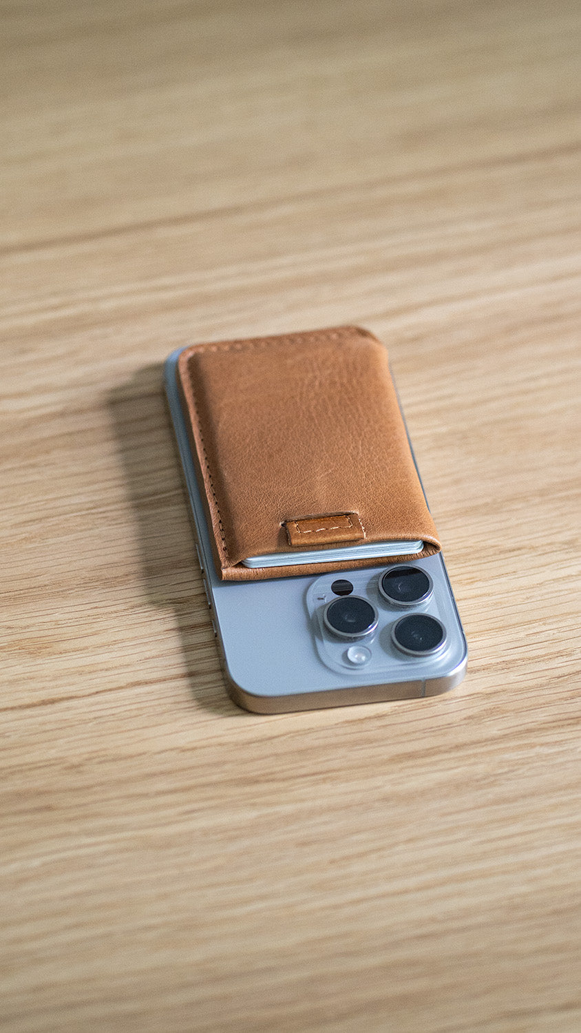 Brown leather card wallet placed on the back of an iPhone on a wooden table.