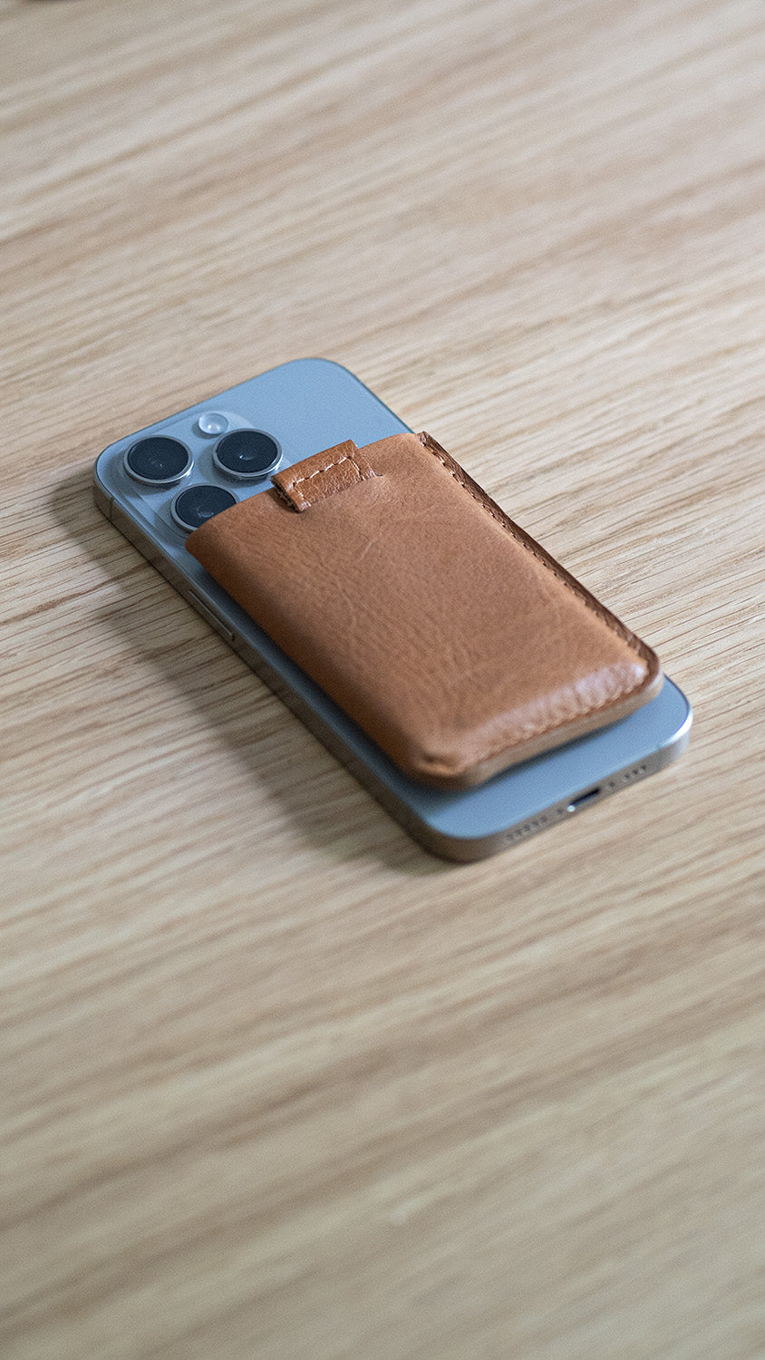 Brown leather magsafe wallet attached to the back of an iPhone on a wooden surface.