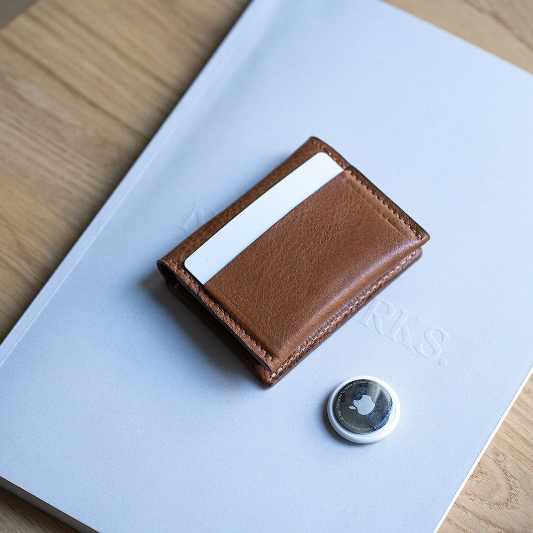 Brown leather slim card wallet with a front card slot, placed on a book next to an Apple AirTag tracker on a wooden table