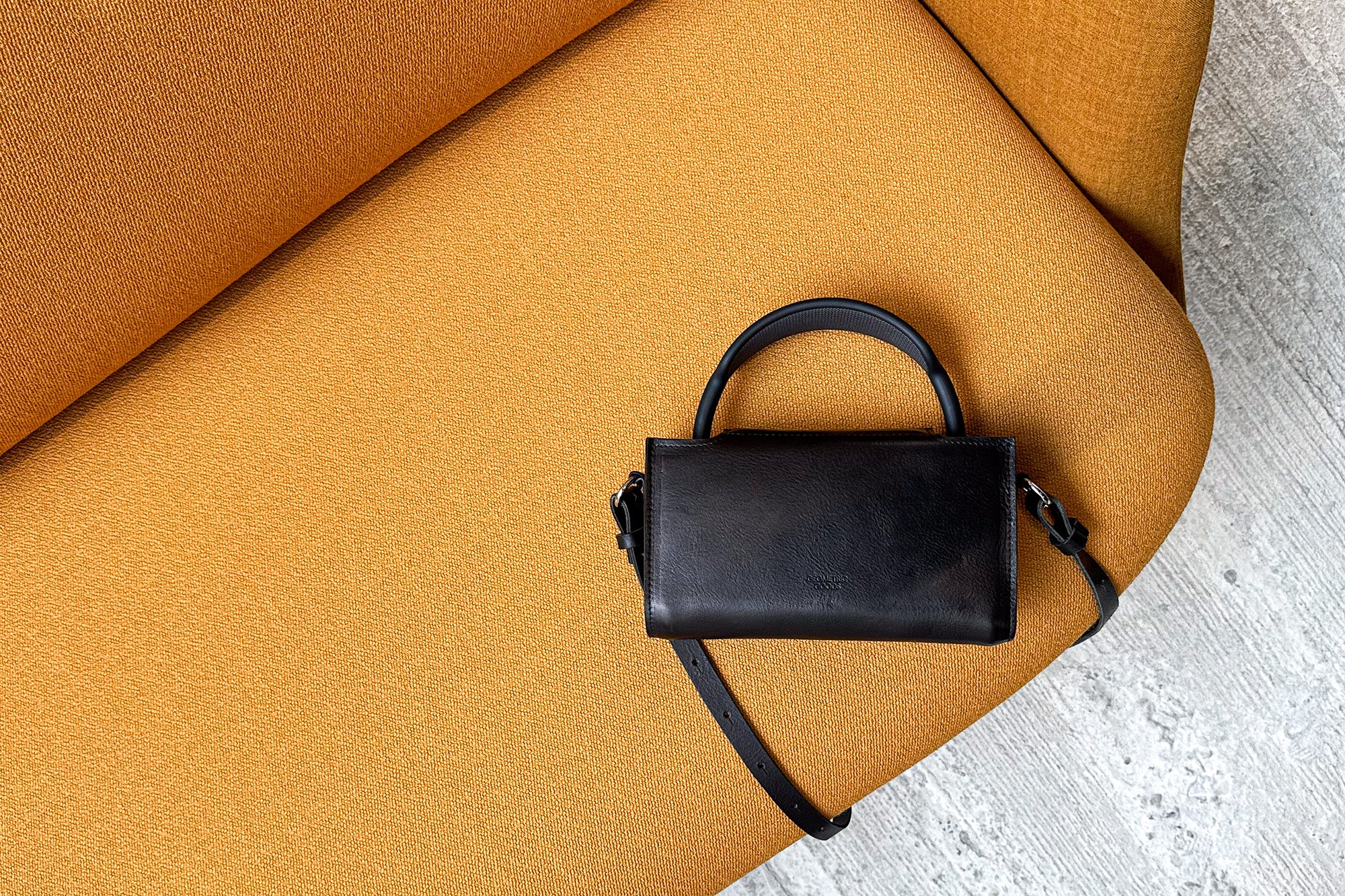 Black leather AirPods Max case bag with shoulder strap and handle, photographed on a mustard-yellow sofa (top-down lifestyle shot)