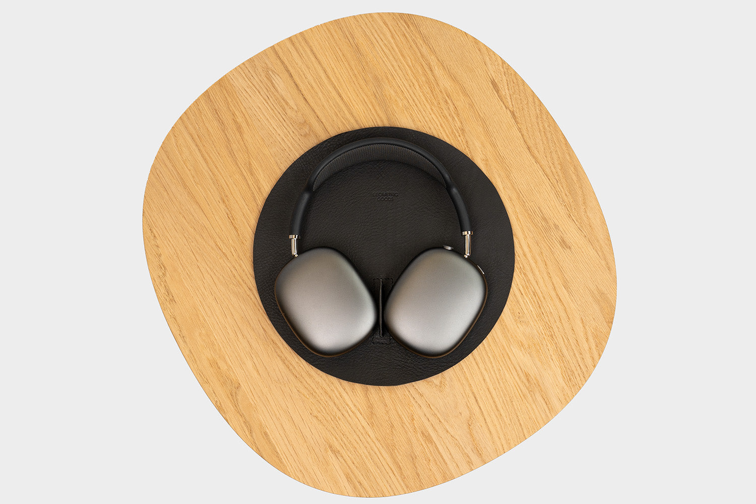 Top-down view of space-gray AirPods Max resting on a round black leather stand with a small center divider, placed on a light oak tabletop.
