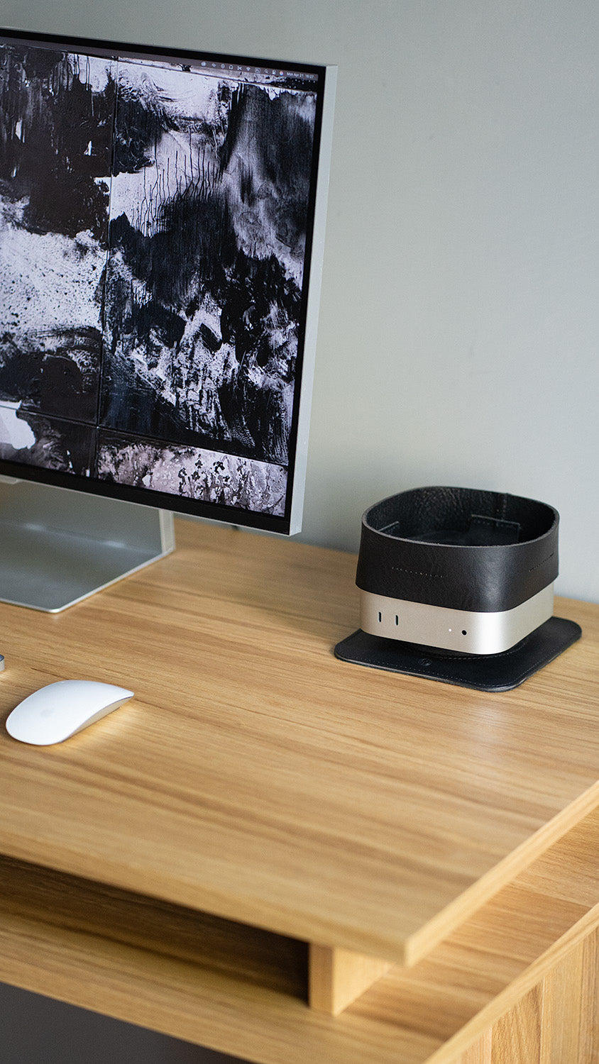 Mac mini desktop computer with a black leather tray placed on top, on a wooden desk beside a monitor