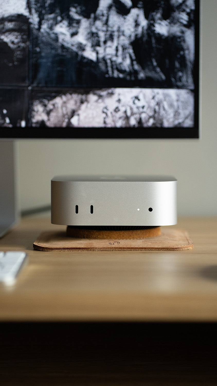 mac mini computer resting on a leather stand on a wooden desk, viewed from the front with a monitor in the background