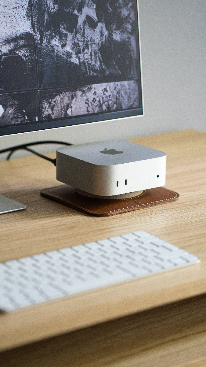 Small desktop computer resting on a leather stand on a wooden desk, with monitor and keyboard in the background