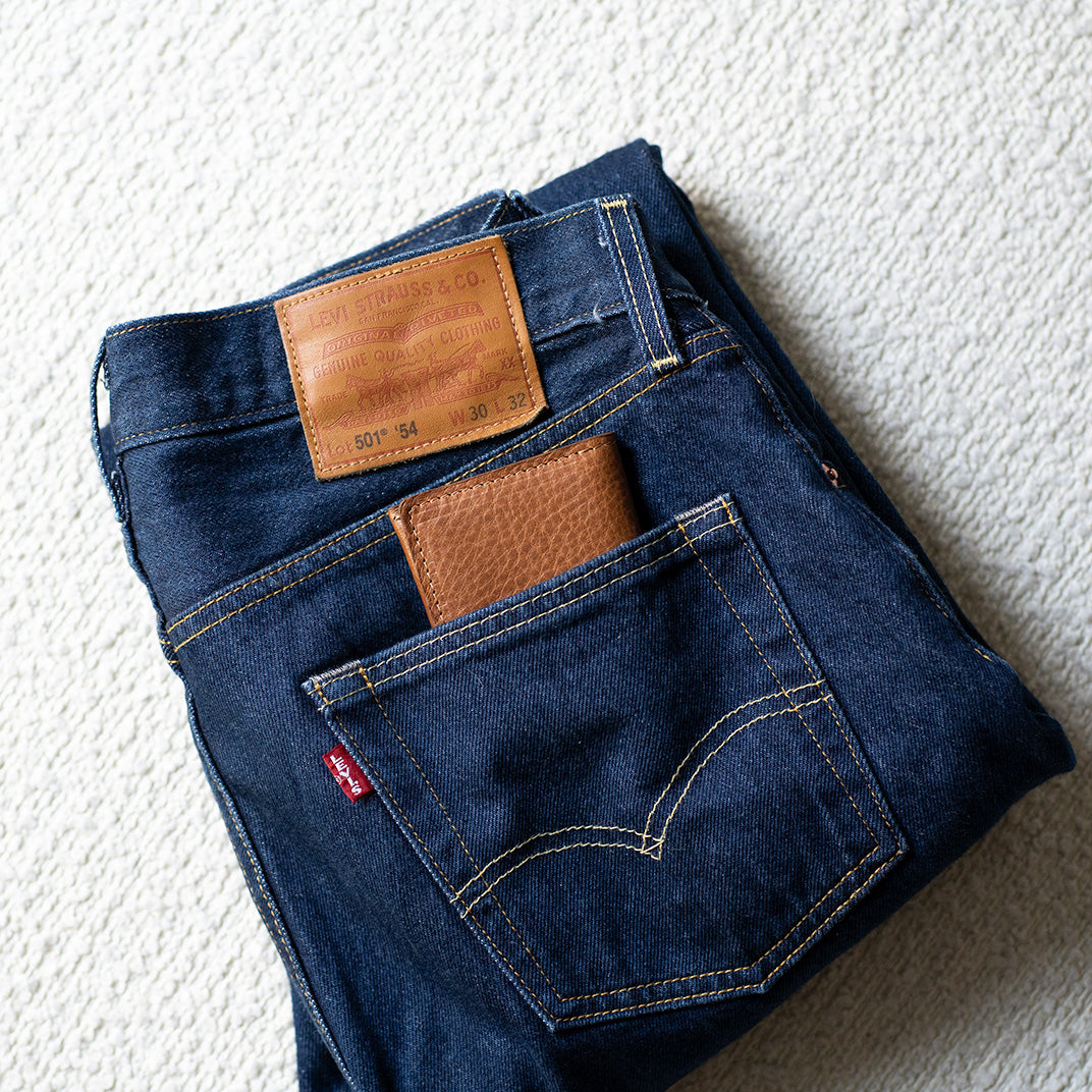 Brown leather slim wallet partially tucked into the back pocket of blue jeans, showing a compact everyday carry fit