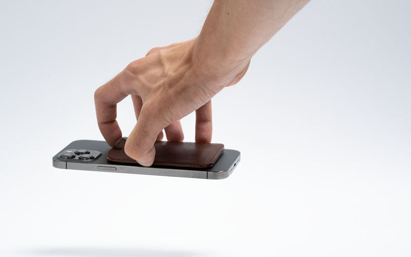 Hand lifting a leather card wallet magnetically attached to the back of a smartphone.