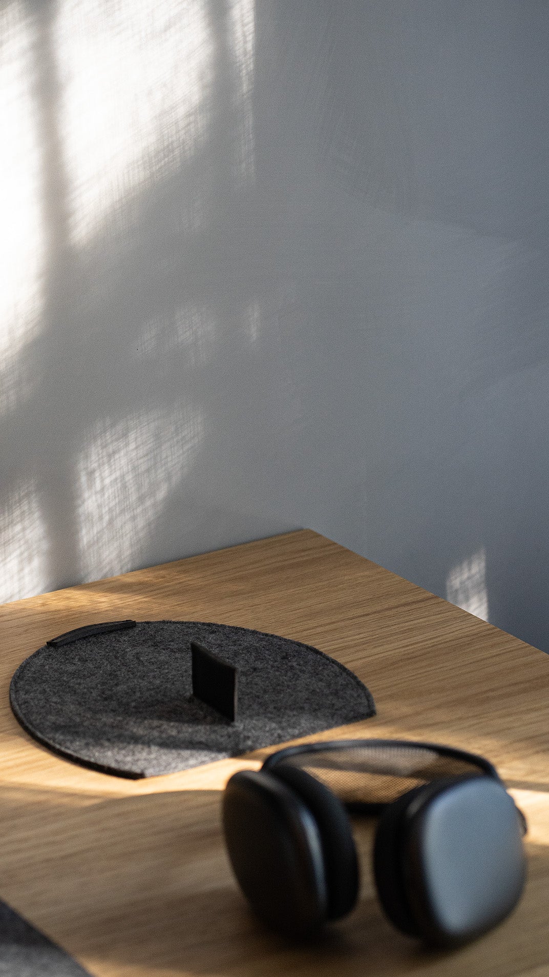 Sunlit oak table with soft window-shadow patterns; round dark gray wool-felt pad with a vertical slot/stand insert, and black AirPods Max headphones blurred in the foreground